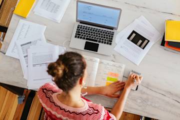 woman working on a desk with a laptop and several document and taking some notes.