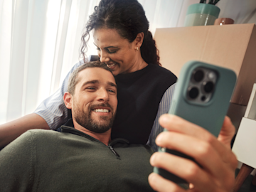Una pareja sonriente mira un smartphone en primer plano, con cajas de mudanza visibles detrás de ellos.