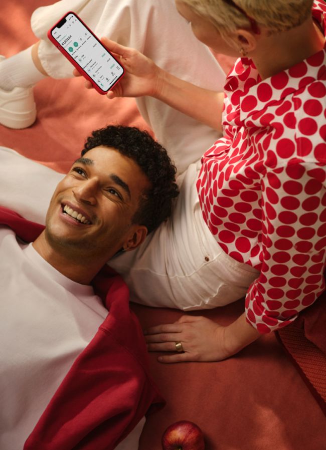 Two youngsters are relaxing on a blanket using the smartphone with N26 app on it while smiling.