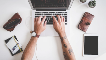 Un uomo con le braccia tatuate che scrive al computer su un tavolo bianco con occhiali da sole, portafoglio, taccuino e iPad.