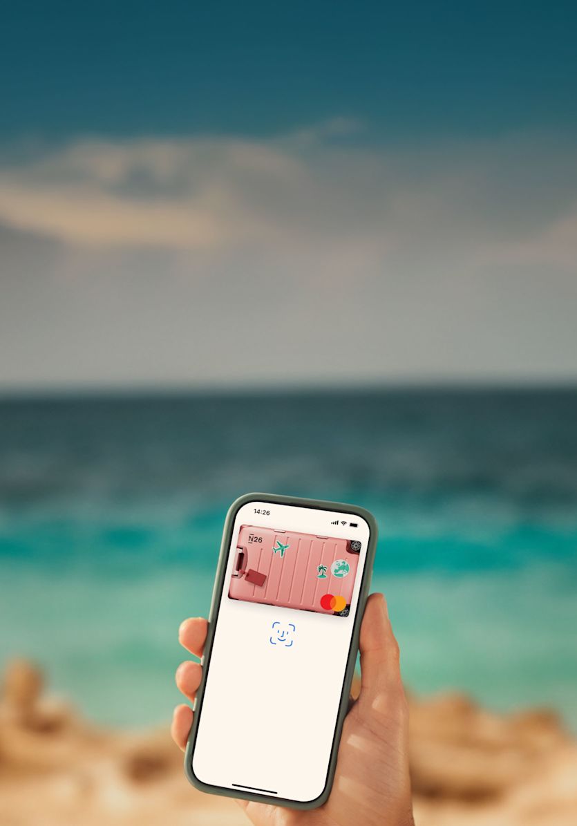 A person is holding a smartphone near the beach, with the ocean and sand blurred in the background. On the phone screen, a digital N26 card with a pink suitcase design is displayed in a mobile wallet app, ready for contactless payment. The screen also shows the Face ID icon, indicating authentication is in progress. The scene highlights modern, convenient mobile payments while traveling.