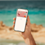 Une main tient un smartphone sur une plage, affichant une Mastercard N26 numérique rose à l'écran.