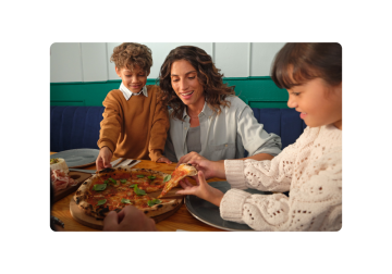 Una famiglia sta gustando una pizza insieme in un ristorante. Il membro più giovane della famiglia sta prendendo una fetta mentre suo fratello e sua madre osservano.
