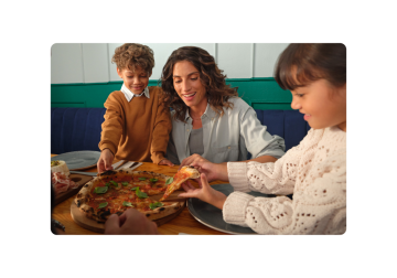 Una familia está disfrutando de una pizza juntos en un restaurante. El miembro más joven de la familia está alcanzando una porción mientras su hermano y su madre observan.