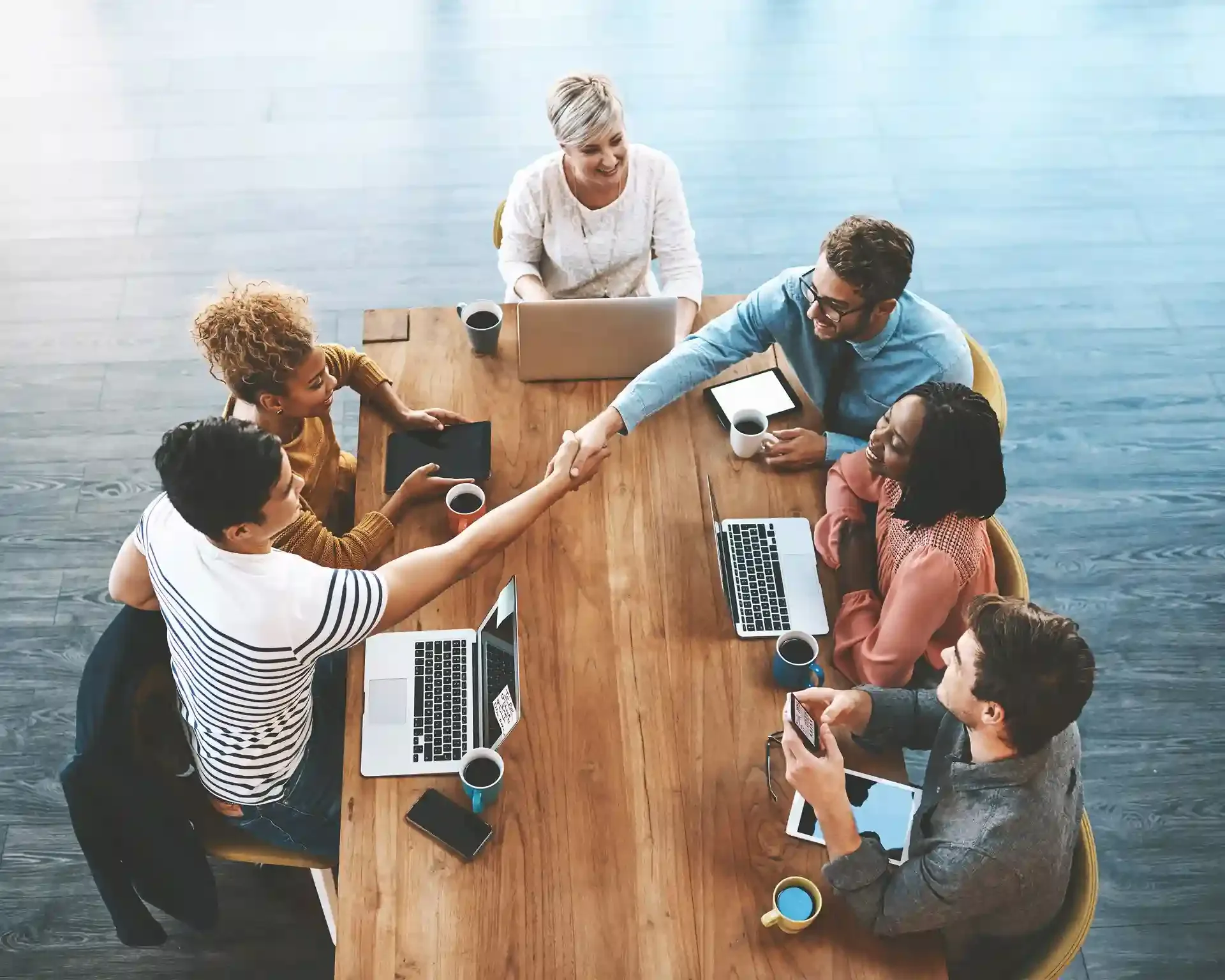 Two coworkers shake hands across a table during a team meeting with laptops and coffee.