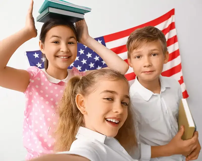 Three smiling children posing in front of an American flag, one holding books on her head.