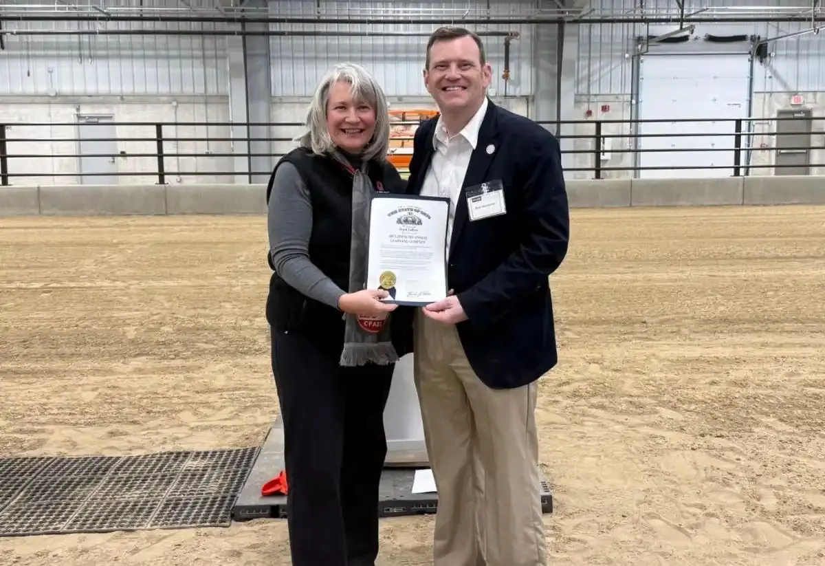 Two people smiling and holding a framed certificate inside an indoor arena.
