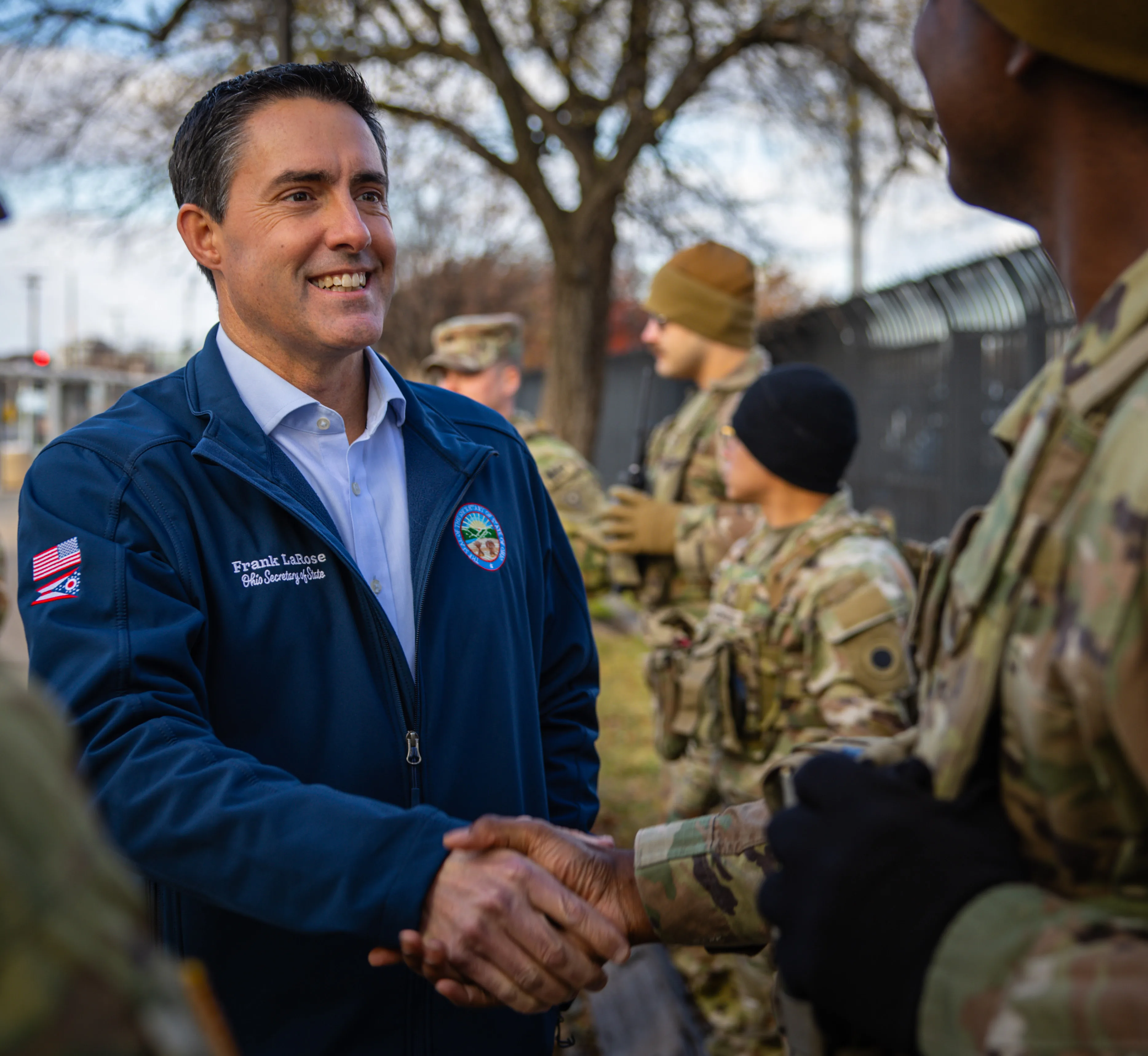Man in a jacket shaking hands with a service member in uniform while other soldiers stand nearby outdoors.