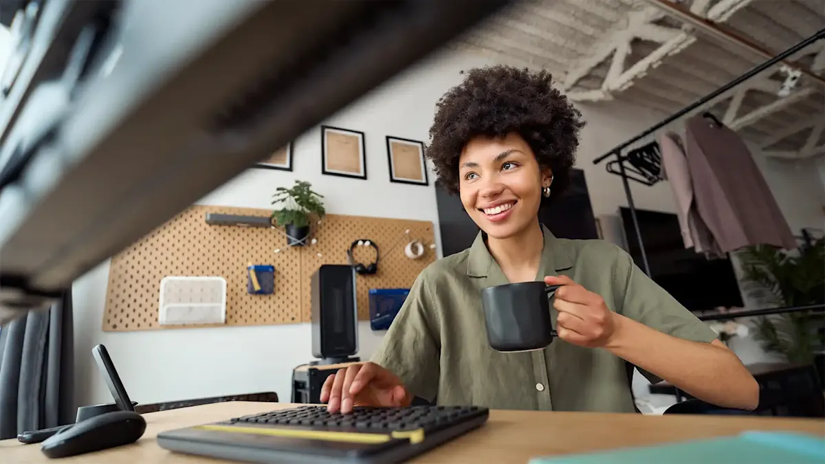 Woman smiling at a desk while typing on a keyboard and holding a coffee mug.