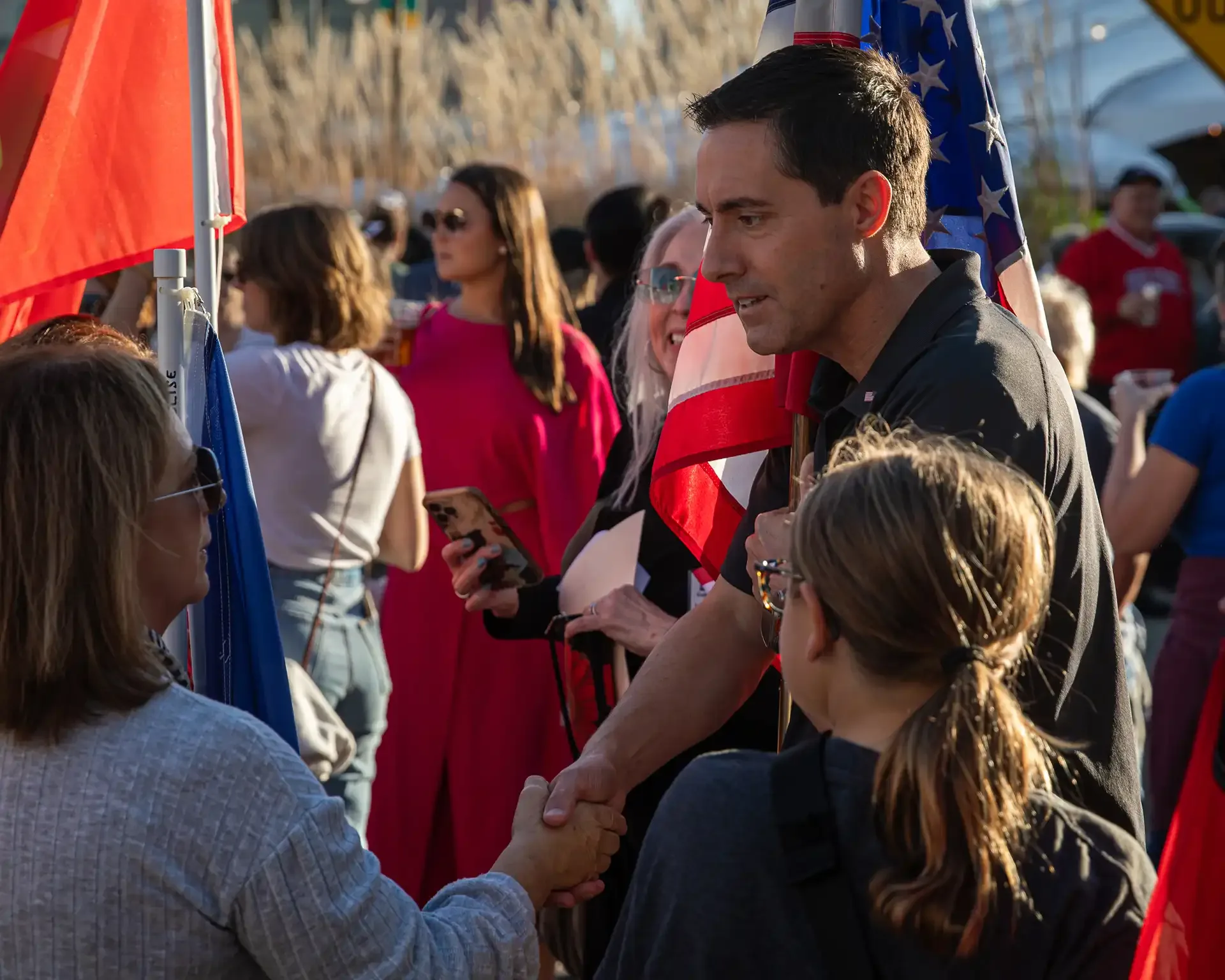 Man shaking hands with attendee at outdoor event with American flags.