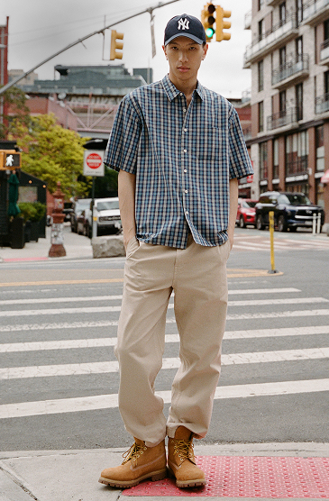 Man wearing NY Yankees cap, blue plaid shirt, and beige pants stands on urban crosswalk with buildings and traffic lights behind.