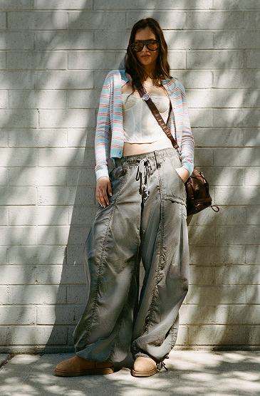 Woman in sunglasses wearing striped cardigan, white top, and oversized cargo pants with crossbody bag against white brick wall.