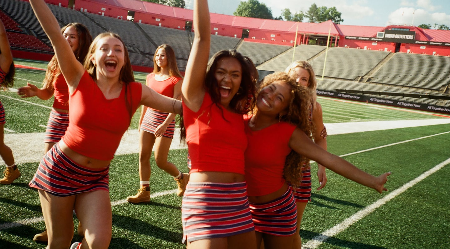 Group of young people in matching white pants and casual tops standing on a football field under blue sky with clouds.