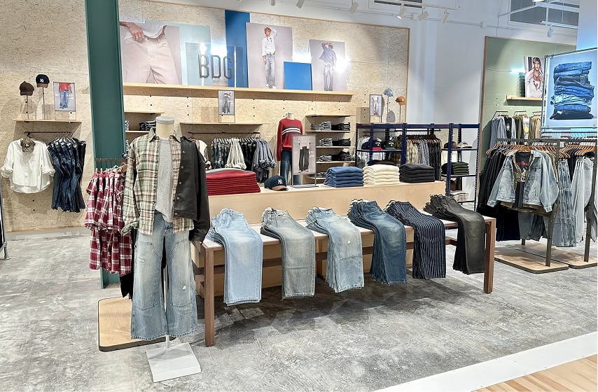 Modern clothing store interior with folded jeans displayed on wooden table, shirts on racks, and fashion photos on walls.