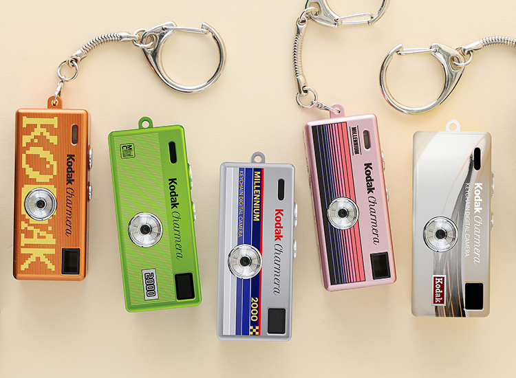 Five colorful Kodak camera-shaped keychains in orange, green, blue, pink, and white arranged on a beige background.