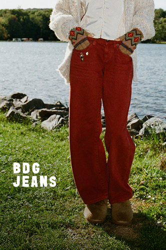 Woman in white textured cardigan and black wide-leg jeans standing by lakeside with green grass and blue sky background.
