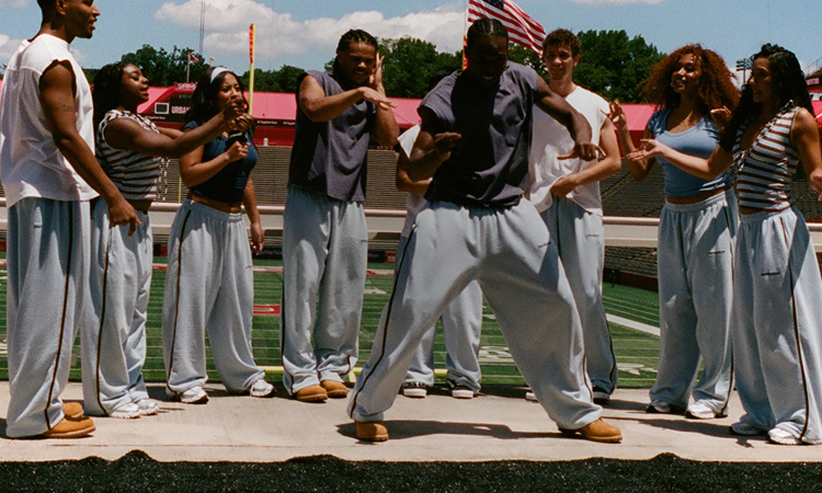 Group of diverse young people in casual athletic wear standing together on a football field with stadium seating in background.