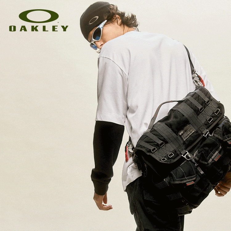 Man wearing Oakley sunglasses, black cap, white shirt, black shorts, and carrying a large black tactical backpack.