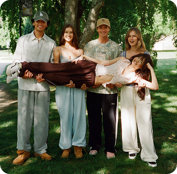 Four people holding a woman horizontally in a trust fall pose outdoors on a sunny lawn with trees in the background.