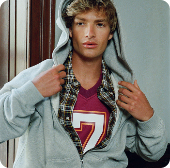 Young man in gray hoodie over plaid shirt and red graphic tee, posing by window with neutral expression.