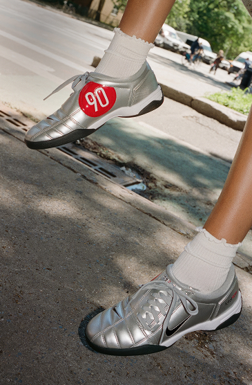 Close-up of legs wearing white socks and silver Nike sneakers with red number 90 patch, standing on concrete steps outdoors.