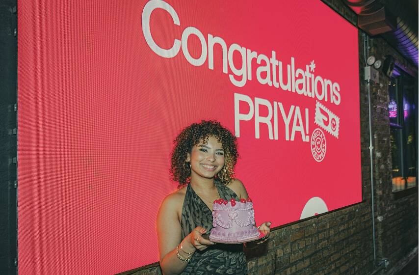 Woman in black dress holding purple cake stands in front of large red digital screen displaying "Congratulations PRIYA!"
