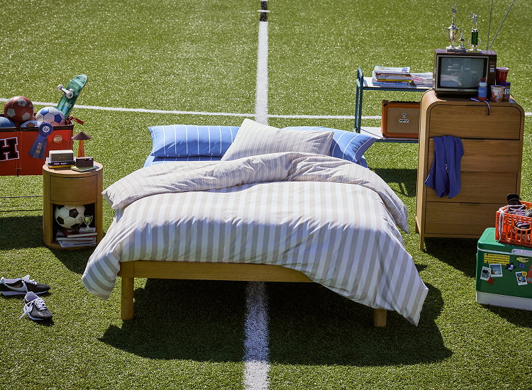 Bed with blue and white striped bedding on artificial turf football field with sports equipment and coolers nearby.