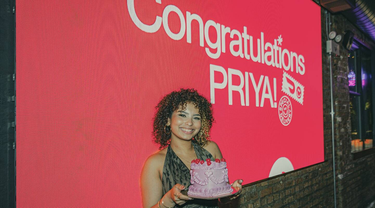 Woman with curly hair holding pink celebration cake in front of large red digital screen displaying congratulations message.