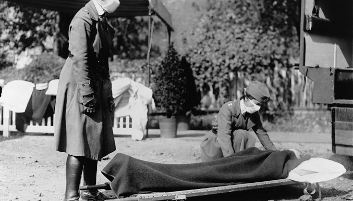 Photo from 1918 of 2 masked nurses outside an ambulance station demonstrating how to care for a pandemic patient on a stretcher.