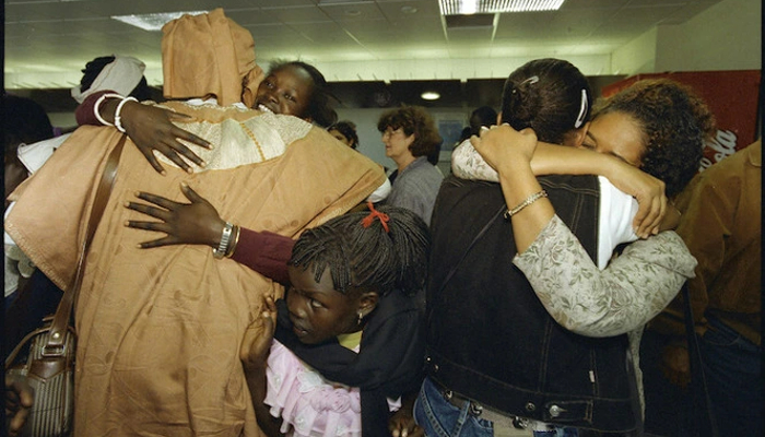 Colour photo of 2 pairs of people hugging. A young girl squeezes through between them. Other people are in the background.