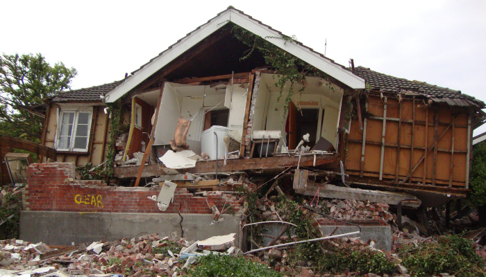 Colour photo of a house severely damaged by the 2011 Christchurch earthquake. A pile of debris lies in front of the house. 