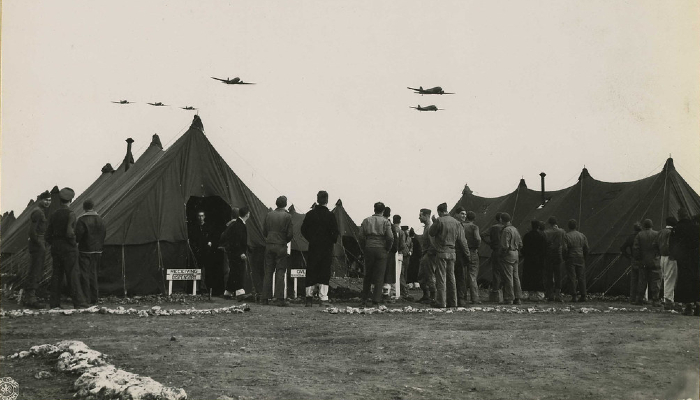 1942 black and white photo of people standing outside an evacuation hospital, watching warplanes fly over.