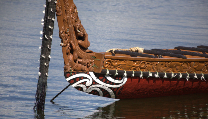 Colour photo of a waka (canoe) showing the taurapa (stern post) and kei (stern) decorated with carvings and kōwhaiwhai (painted scroll ornamentation) patterns.