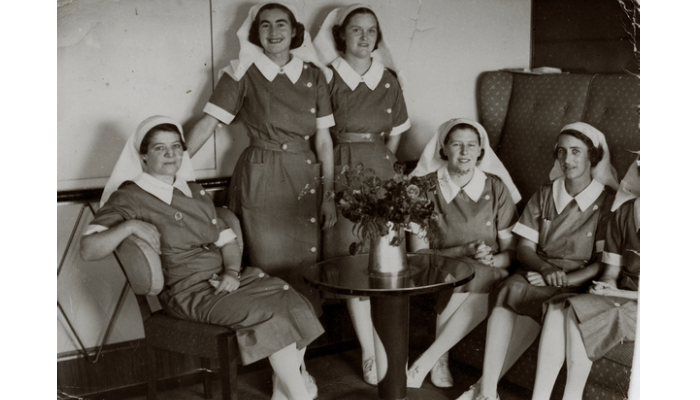 Black and white photo from WW2 of five nurses wearing uniforms. The two in the middle are standing. The others are sitting.