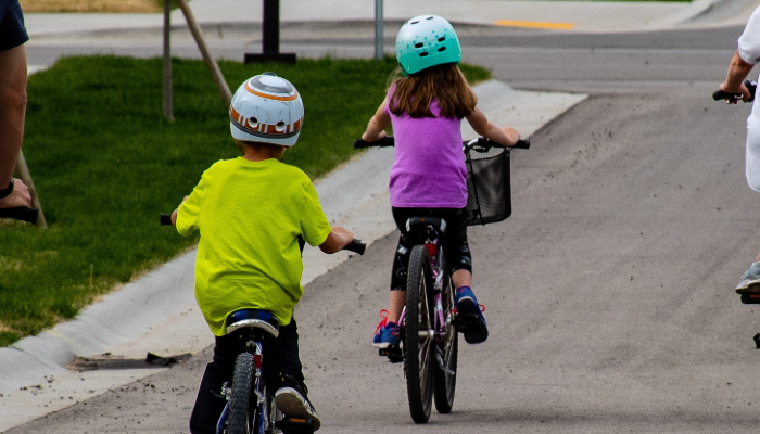 Colour photo of 2 children riding cycles. Both children wear safety helmets, bright-coloured tops and fitted trousers. 