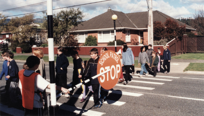 1999 colour photo of a group of primary school children using a pedestrian crossing. One studnet wears an orange vest and holds out a sign saying "School Patrol. Stop."