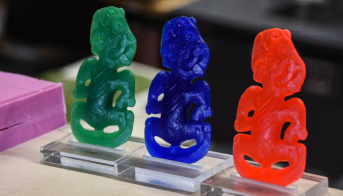 Colour photo of 3 hei tikis (carved pendants in human form) in a row. One is green, one is blue, and one is red.