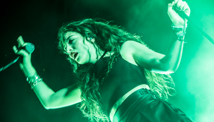 Photo of Aotearoa NZ singer Lorde wearing a sleeveless crop top and high-waisted trousers. She holds a mike in one hand.