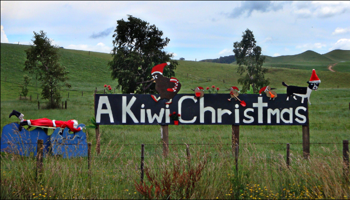 Colour photo of a sign in a field in Aotearoa New Zealand with the words 'A kiwi Christmas'. The sign is decorated with kiwis and a dog wearing Santa hats.