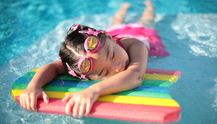 Colour photo of a girl resting on a rainbow coloured paddle board in a swimming pool.