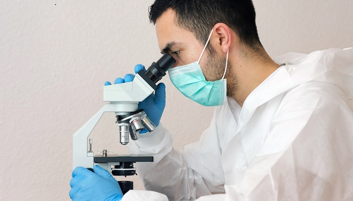 Colour photo of a scientist looking into a microscope. They are wearing PPE — including a mask, gown and gloves.