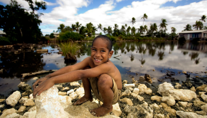 Colour photo of a smiling child playing on rocks in Nuku'alofa, Tonga. There is floodwater in the background.