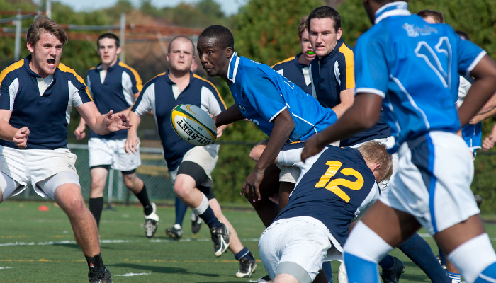 Colour photo of 2 rugby teams playing in a field. One of the players is holding a rugby ball. 