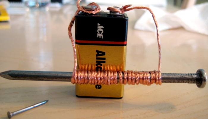 Colour photo of an electromagnet. It shows a copper wire wound around a nail. The ends of the wire are attached to the terminals of a battery.