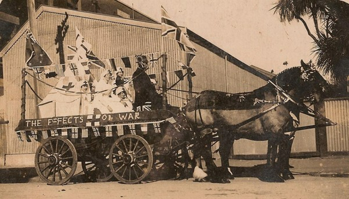 1918 black and white photo showing a wagon pulled by horses, decorated with flags. People in nurse uniforms are in the wagon. There is a banner on the side which reads 'The effects of war'.