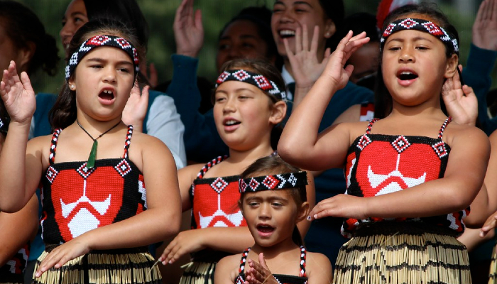 2012 colour photo of a group of students in piupiu (skirt), pari (bodice) and tīpare (headband) performing a waiata ā-ringa (action song).