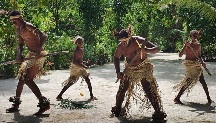 Colour photo of four ni-Vanuatu people performing on sand wearing woven garments around waists and surrounded by trees.