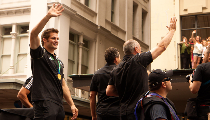 Colour photo of Richie McCaw waving to people on a street after winning the 2011 Rugby World Cup. He wears his winning medal on his neck.