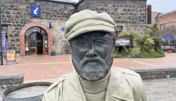 Close up photo of a statue of Captain Henry Cain. Behind the statue is the Landing Service Building on George Street, Timaru.