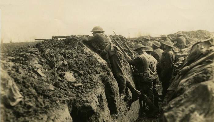 1918 black and white photo showing Aotearoa NZ soldiers in a trench in the Somme region during WW1. One of the soldiers is pointing his gun over the side of the trench.